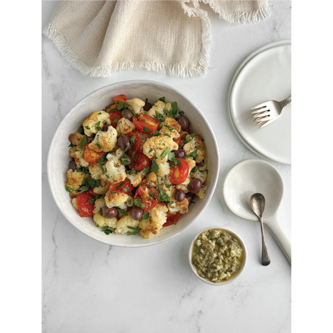 Photograph taken from above of a bowl of pan-fried cauliflower, roasted tomatoes, Niçoise olives, and a ramekin of lemon caper sauce